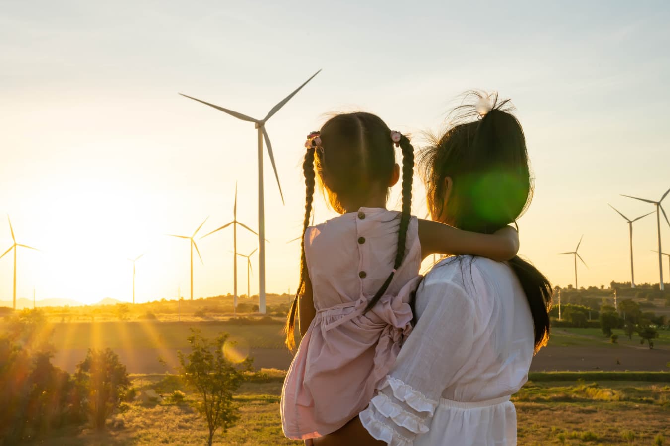 Mutter blickt mit der Tochter auf dem Arm in den Sonnenuntergang hinter einem Windpark